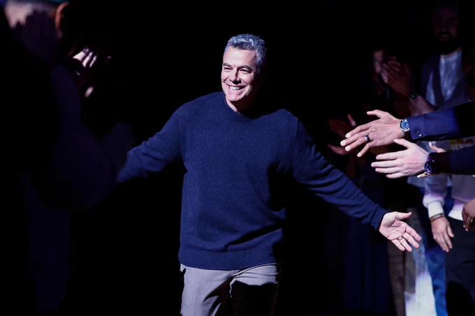 Tony Bennett wearing a sweater and pants is smiling with arms outstretched, surrounded by people clapping in a dimly lit setting.