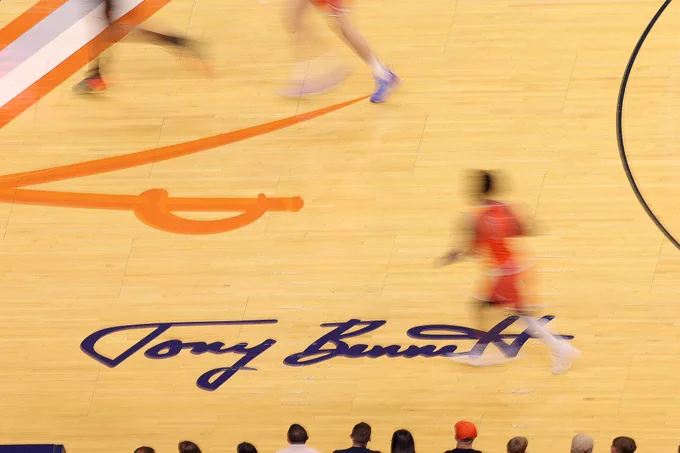 A basketball court with blurred players in motion. "Tony Bennett" is signed on the court's wooden floor. Spectators are seated at the bottom edge of the image.