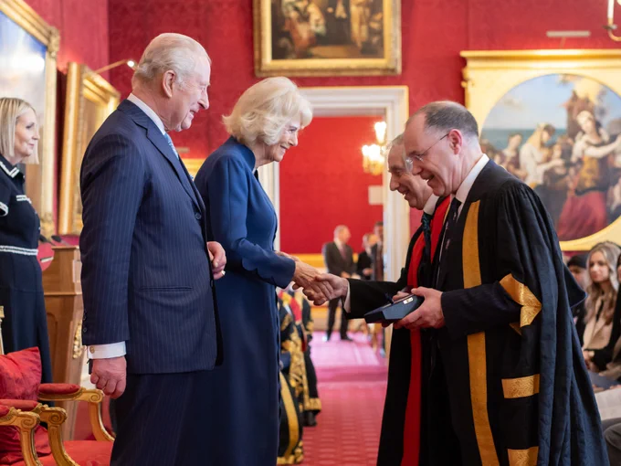 Professor William Powrie and Vice-Chancellor Professor Mark E. Smith stand in a grand room with red walls and ornate paintings. The King and Queen greet them, with the Queen shaking Professor Powrie’s hand while the Vice-Chancellor holds the award. Guests are seated in the background.