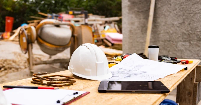 A construction site with a table holding a hard hat, blueprints, a tablet, coffee cup, and various tools, with building materials in the background.