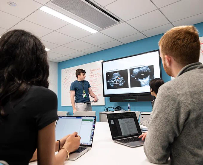 A person stands at the front of a classroom, presenting ultrasound images on a screen. Several individuals are seated at a table with laptops, taking notes.