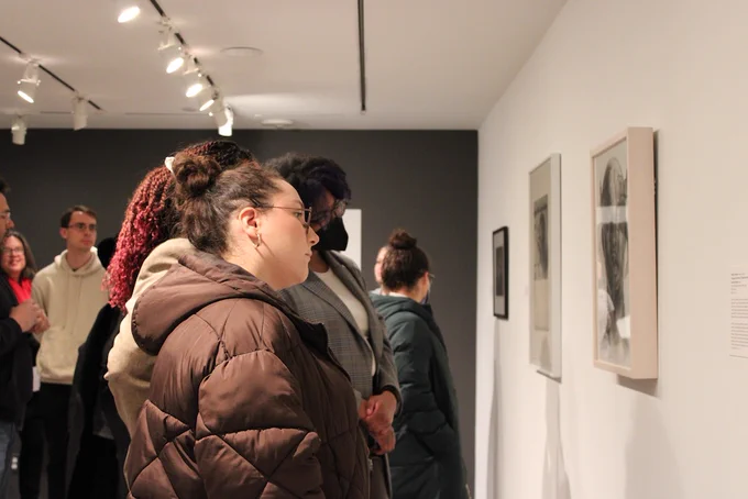 A group of people in the University Museum of Contemporary Art at UMass Amherst are observing framed art on a white wall. Several spotlights illuminate the artwork. The room has a dark grey accent wall in the background.