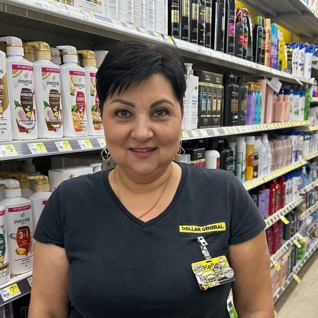 A woman with dark hair smiles in front of shelves of hair products. She wears a black shirt, a DOLLAR GENERAL name tag that says, \"text@1,\" and a gold chain. Products like PANTENE and Dove can be seen on the shelves behind her.