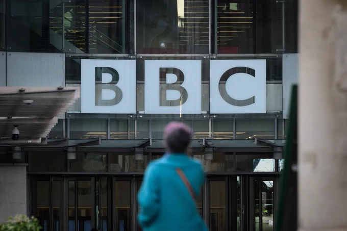 The BBC logo on the front of a building, with a member of the public walking in the foreground.