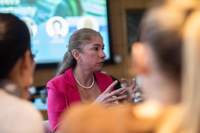 A woman in a pink blazer, wearing pearl jewelry, gestures while speaking in a meeting. The setting is professional, with blurred attendees in the foreground.