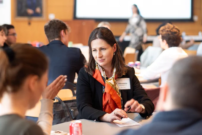 A woman in business attire, wearing a name tag, sits at a table engaged in discussion during a meeting. Others in the background create a collaborative atmosphere.
