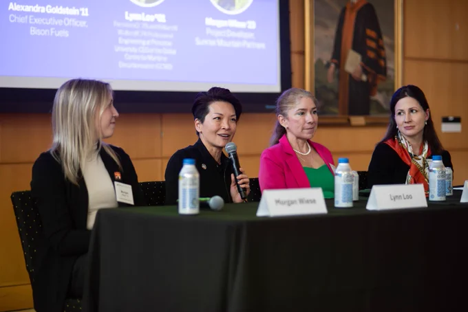 Four women sit at a conference table in a panel discussion. One speaks into a microphone while others listen attentively.