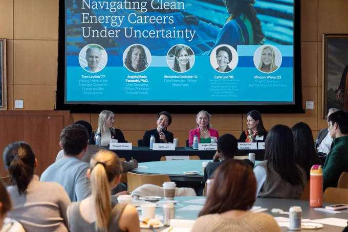 A panel of five speakers, seated at a table, discusses clean energy careers. The backdrop features a large screen displaying speaker info and the event title, "Navigating Clean Energy Careers Under Uncertainty." 