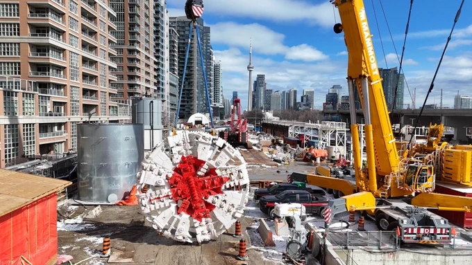 Construction site in an urban area with a large tunnel boring machine in the center. Surrounding high-rise buildings suggest a city environment. A yellow crane is on the right, and the CN Tower is visible in the background. Construction materials and vehicles are scattered around the site. 