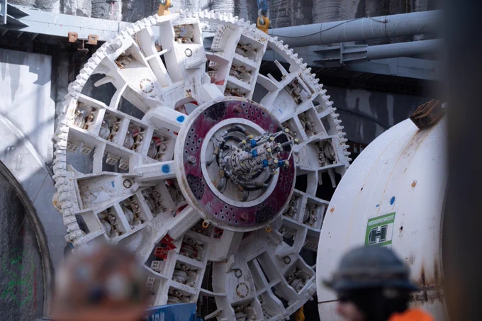 A large tunnel boring machine is positioned at a construction site, with its round, segmented cutter head prominently visible. Two workers in hard hats are in the foreground, slightly blurred. The background shows concrete walls and metal beams. 