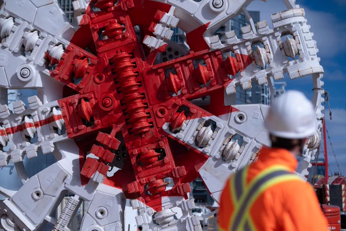 A large, intricate tunnel boring machine with red and white components is displayed prominently. A person wearing a white hard hat and an orange high-visibility jacket is visible in the foreground. The sky is clear and blue. 