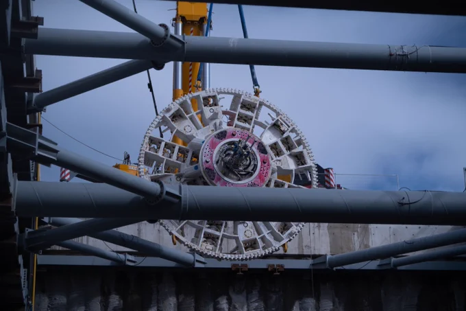 A large, intricate tunnel boring machine with red and white components is displayed prominently. A person wearing a white hard hat and an orange high-visibility jacket is visible in the foreground. The sky is clear and blue. 