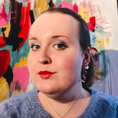 A woman standing in front of abstract art with her hair pulled back, sparkly blue eyeshadow, and red lipstick looks confidently into the camera. 