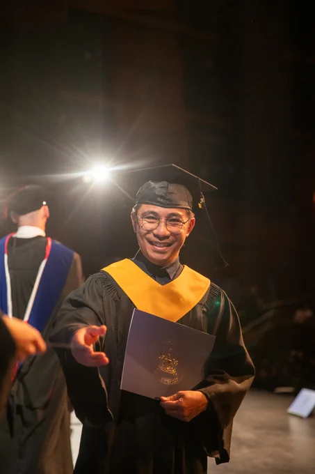 A graduate in a cap and gown smiles, holding their credential on stage during the ceremony, with bright lights in the background.