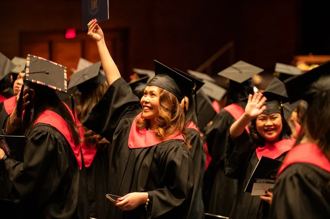 A group of graduates in black gowns and mortarboards with red stoles celebrate in a ceremony. One person is holding up a diploma, while others smile and wave.