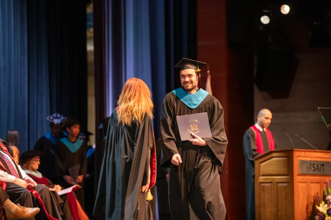 A graduate is receiving a credential on stage in a ceremony setting, with a blue curtain in the background. 