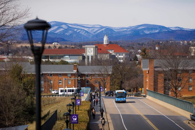 Snow-covered blue ridge mountains rise behind JMU's campus as students walk over the I-81 bridge.