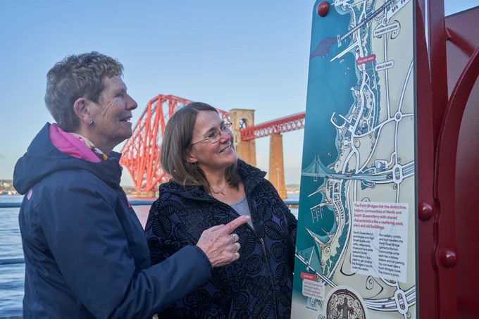 Two women looking at a tourist map of the Forth Bridges Trail in South Queensferry with the river Forth and Forth Bridge behind. 