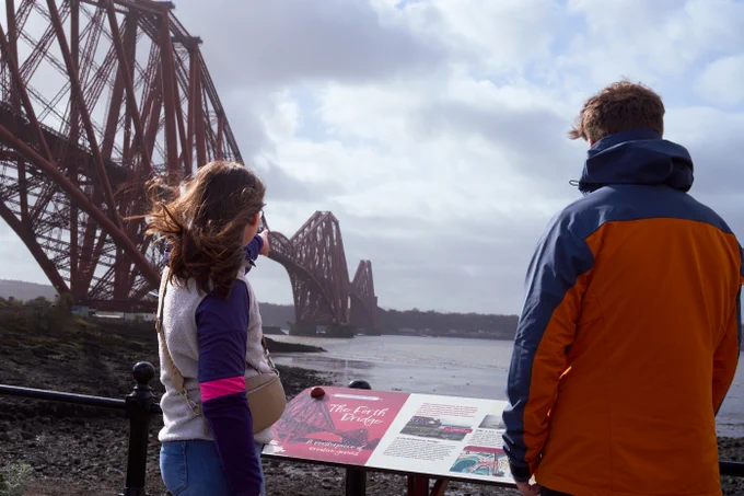 Man and woman in warm clothes looking at the Forth Bridge in North Queensferry with a Forth Bridges Trail sign in front of them.