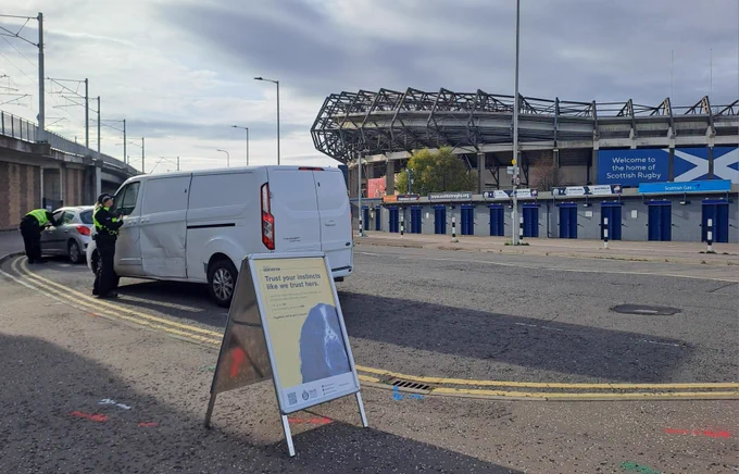 Project Servator police officers standing beside a white van and silver car on a street near Murrayfield Stadium in Edinburgh. A yellow Project Servator information sign is positioned in the foreground, with the stadium visible in the background.