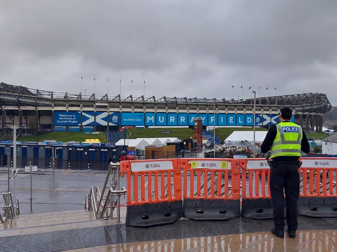 A Project Servator police officer wearing a high-visibility vest looks out over a cordoned area at Murrayfield Stadium in Edinburgh. Temporary barriers are visible, with stadium signage in the background.