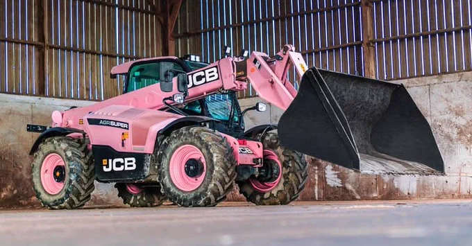 A pink JCB Agri-Super loader, with black bucket, is parked inside of a barn. The JCB logo is visible on the side of the vehicle, and light streams in through the barn's walls.