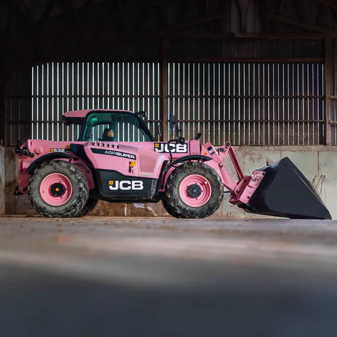A pink JCB Agri Super Loadall inside a barn, the tractor has black accents and wheels. A person is visible in the driver's seat. The barn has a wood structure with vertical white slats in the back.