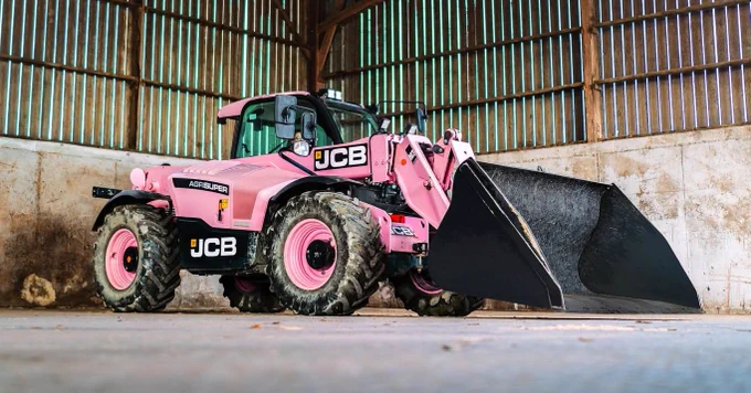 A pink JCB telehandler with a black bucket parked inside a shed with a wooden roof and vertical plank walls. The tractor's tires are muddy, and the JCB logo is visible on the side.