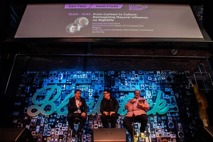 Three speakers sit on stools on a stage in front of a large wall of speakers, taking part in a panel discussion. Above them, a large screen displays the event session details titled “From Curfews to Culture: Reimagining Mayoral Influence on Nightlife.”