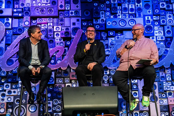 Three speakers sit on high stools on a stage, each holding a microphone during a panel discussion. They are positioned in front of a large backdrop made from stacked speakers in various shapes and sizes, creating a vibrant, music‑themed setting.