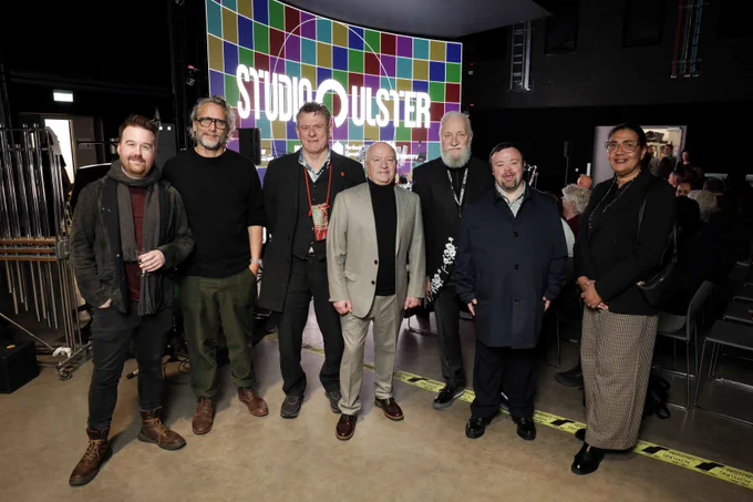 Seven people stand indoors in front of a large screen displaying a colourful Studio Ulster sign. From left to right are Michael Weir Director of RENDR Festival Fede Álvarez writer producer and director Andrew Kötting artist and filmmaker Professor Frank Lyons Director of CoSTAR Screen Lab Professor Paul Moore Director of Future Screens NI Dr James Martin MBE actor and Sophia Alexandersson Chief Executive of ShareMusic and Performing Arts.