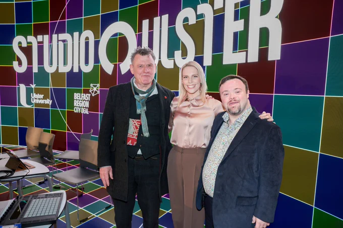 Three people stand together in front of a colourful tiled wall with the words Studio Ulster displayed behind them. From left to right are Andrew Kötting artist and filmmaker Shelley Cowan Disability Entrepreneur and Accessibility Consultant and Dr James Martin MBE actor.