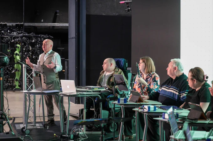 Professor Frank Lyons stands at the front of a studio introducing performers from Acoustronic. He wears a vest and presents to a seated group using a laptop and microphones in a professional studio setting.