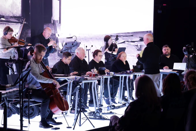Musicians from Acoustronic Hard Rain and Elefantöra perform Zoom Time in a modern ensemble setup. String and wind players are seated under bright stage lighting in a contemporary performance space.