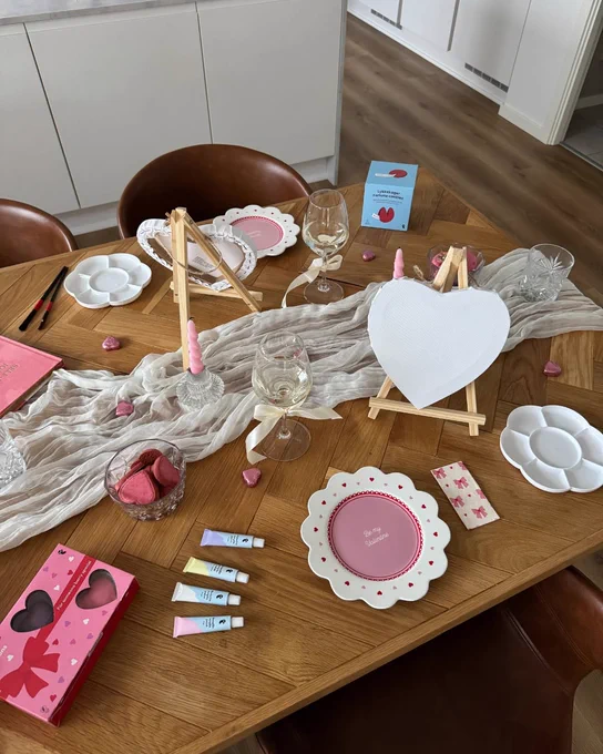 A wooden table set for a painting activity with heart-shaped canvases on easels. The table is decorated with a light fabric runner, scattered pink heart decorations, and paint supplies. Two glasses and plates with heart motifs are also present. The room has a modern, minimalist design with a wooden floor and white cabinets in the background.
