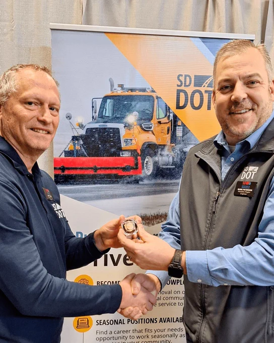 Two men shaking hands while together holding a pin to represent years of service.