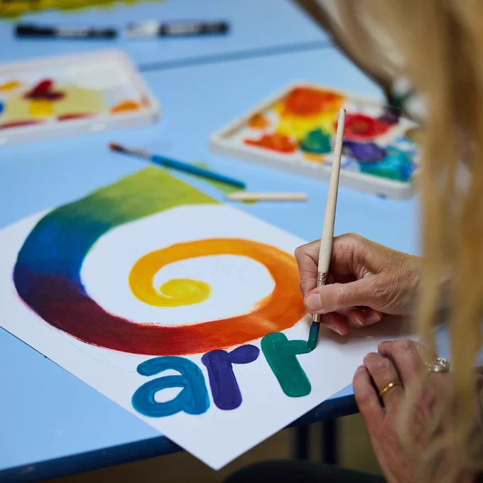 Person painting a colorful spiral and the word 'art' on white paper using a fine brush. Paint brushes and palettes are also scattered across the table.