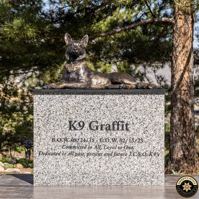 Bronze statue of K9 Graffit lying on a granite memorial pedestal outdoors among pine trees. Pedestal inscription reads: “K9 Graffit” and “B.O.W. 08 24 15 E.O.W. 02 13 23” and “Committed to All, Loyal to One” and “Dedicated to all past, present and future JCSO K9’s.” Jefferson County Sheriff’s Office logo appears in the lower right.
