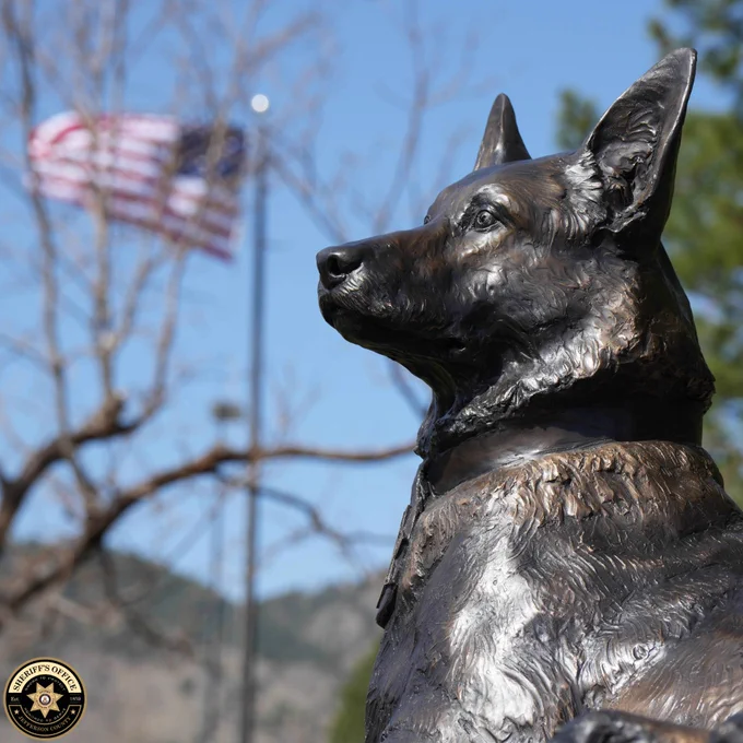 Bronze statue of a K9 Graffit in profile, with a blurred American flag and blue sky in the background. Jefferson County Sheriff’s Office logo appears in the lower left.
