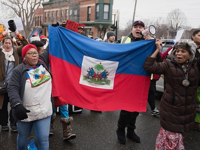Haitian immigrants protest the Trump administration's immigration policies in St. Paul, Minnesota, January 20, 2018. (Fibonacci Blue / CC BY 2.0)  Two Black women are holding a large Haitian flag between them. A white man with a megaphone leads chants, many people are marching behind them. 

See more photos by Fibonacci Blue on Flickr https://www.flickr.com/photos/fibonacciblue/