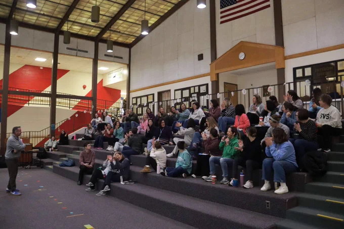 A group of adults seated on tiered bleachers inside a school auditorium listening to a speaker standing nearby.