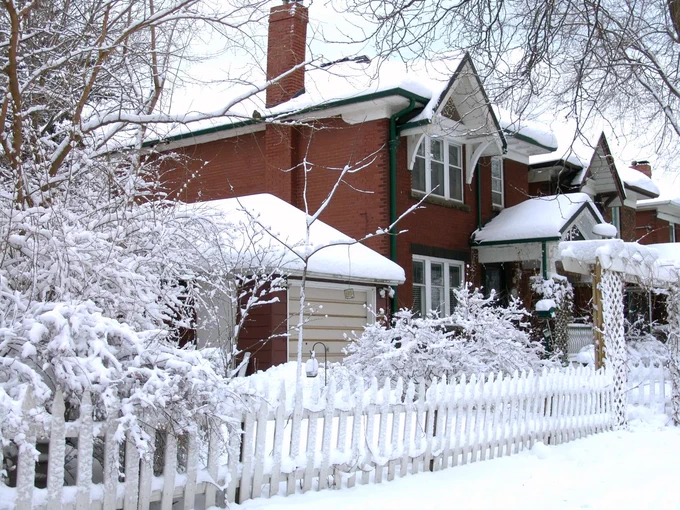 Snow-covered residential area with traditional houses and a white picket fence, trees and shrubs blanketed in snow, creating a picturesque winter scene.