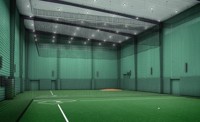 Indoor baseball training facility with green walls and ceiling, featuring a turf field and a pitcher's mound. Nets are suspended from the ceiling, and fluorescent lights provide illumination.