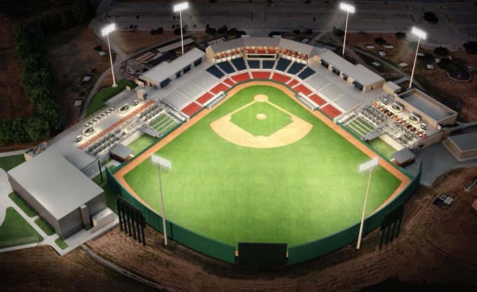 Aerial view of a well-lit baseball stadium at night, showing the field with bases, bleachers, and surrounding areas, including parking and nearby buildings.
