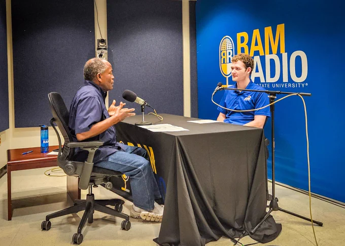 Two individuals sitting in a podcast studio doing a podcast. 