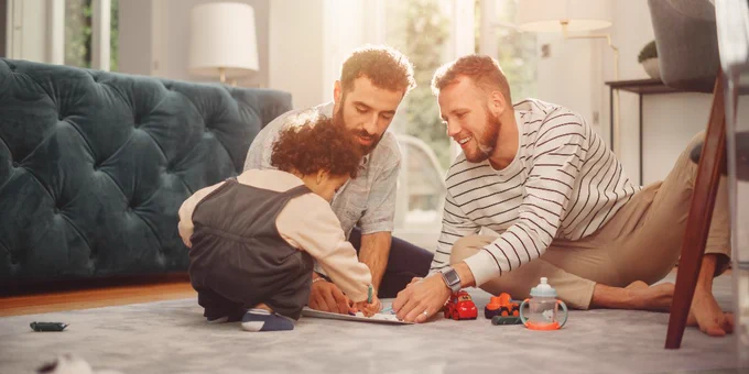 A family (a toddler and two adults) playing on a carpeted floor in a sunlit living room with toys scattered around.