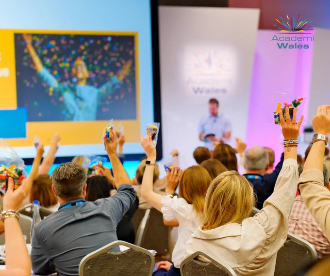 Audience members at an Academi Wales event raising small bags of colorful LEGO bricks during a workshop session.