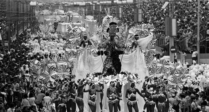 Fotografia em preto e branco registra o desfile da Beija-Flor de Nilópolis no Carnaval de 1978. No centro da imagem, carros alegóricos avançam pela avenida. Em destaque, sobre o primeiro carro, três mulheres com fantasias elaboradas, adornadas por plumas, grandes tecidos e detalhes que reforçam o conceito carnavalesco. À frente e ao redor dos carros, dezenas de componentes desfilam com suas fantasias. Eles usam adereços de cabeça e figurinos bem trabalhados, formando composição coreografada. A avenida está completamente tomada pela multidão.