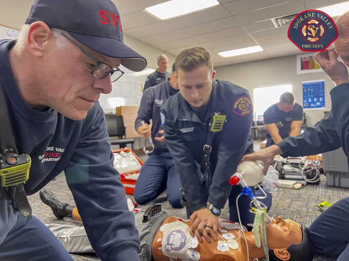 A group of individuals from Spokane Valley Fire Department are engaged in a medical training exercise. They are practicing CPR on a medical dummy, with one person performing chest compressions and another using a bag valve mask. Emergency equipment is visible in the background.