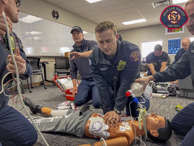 Group of individuals from Spokane Valley Fire Department perform CPR on a medical training mannequin in a classroom setting. Emergency equipment and a whiteboard with notes are visible in the background.
